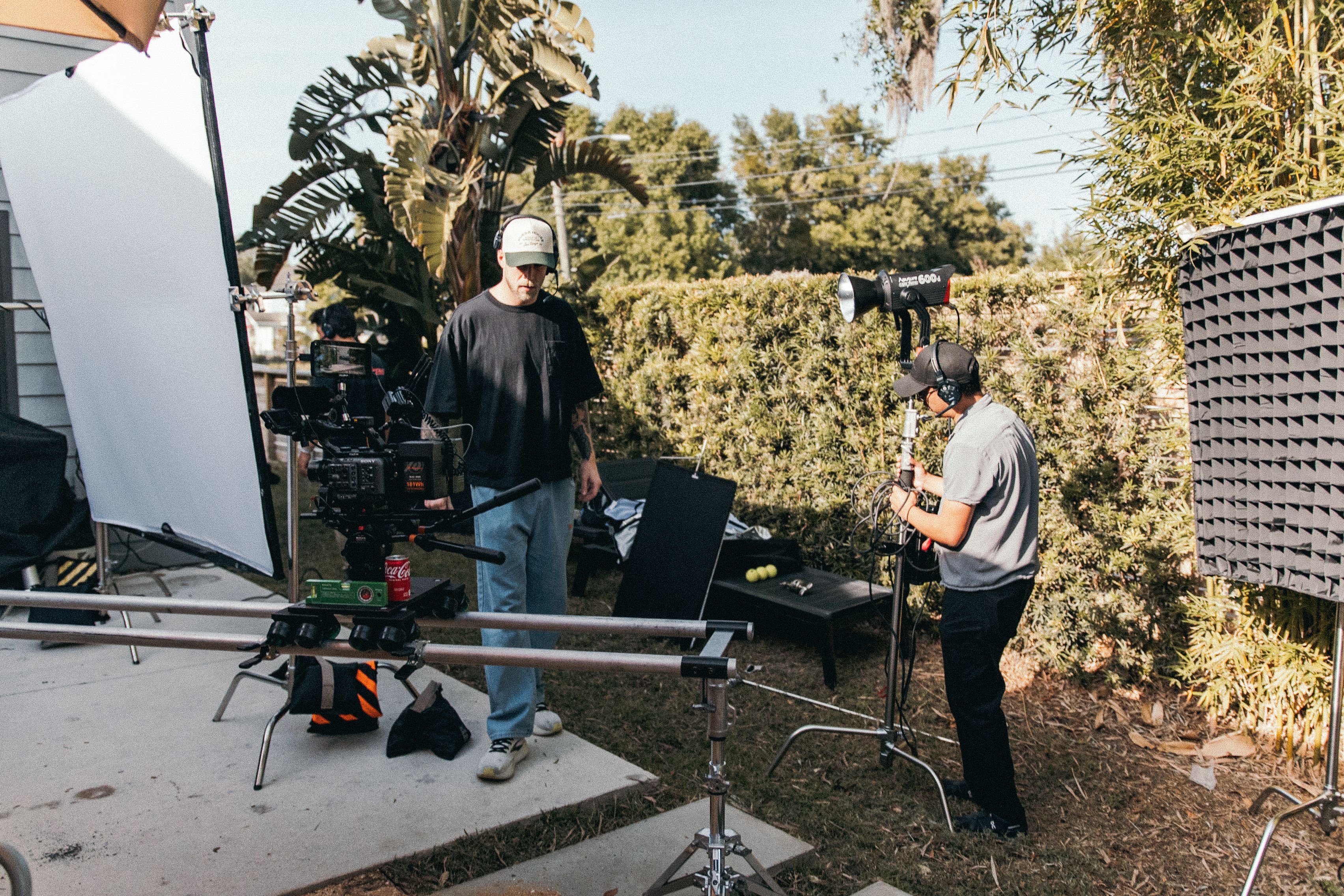 Film crew arranging camera and lighting equipment outdoors beside lush greenery.